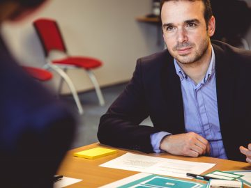 homme assis devant une table avec un coude posé sur la table portant une chemise bleue et une veste de costume sombre