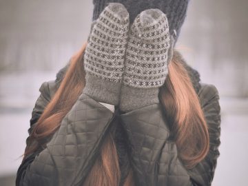 femme aux cheveux longs portant un bonnet foncé une veste kaki et des moufles se cachant le visage