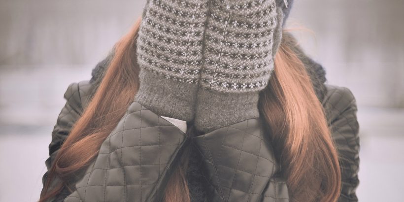 femme aux cheveux longs portant un bonnet foncé une veste kaki et des moufles se cachant le visage