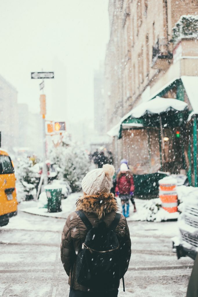 femme dans la rue sous la neige portant un bonnet clair une doudoune et un sac à dos noir